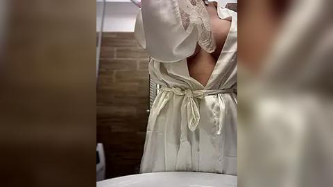 Media: Video of a woman in a satin, off-white robe with lace trim, adjusting her hair in front of a bathroom mirror. Background shows a tiled wall and a blurred white sink.