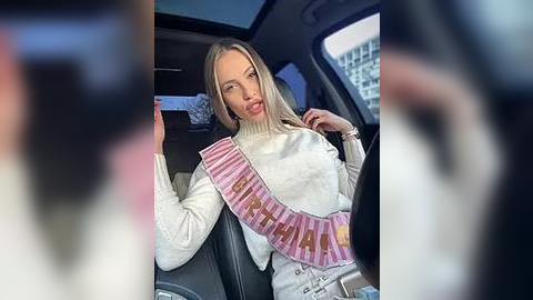 Media: Video of a blonde woman in a white sweater, wearing a pink seatbelt with \"Miss Universe\" printed on it, sitting in the car.