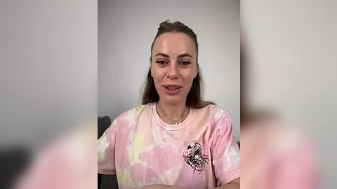 Media: Video of a young woman with fair skin and straight brown hair, wearing a pink tie-dye t-shirt with a skull graphic, seated against a plain grey wall, smiling.