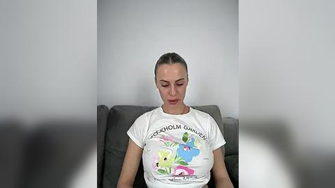 Media: Video of a fair-skinned woman with brown hair tied back, wearing a white T-shirt with a colorful cartoon design, sitting on a gray couch against a plain white wall.