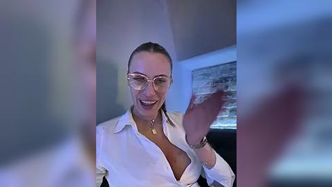 Media: A video of a smiling woman with glasses and an unbuttoned white shirt, her hands moving, blurred figures in the background, a blue-toned indoor setting.