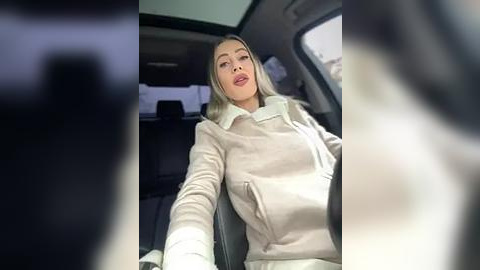 Media: Video of a blonde woman in a beige coat, driving a car with a blurred figure in the foreground, showcasing a modern, well-lit interior.