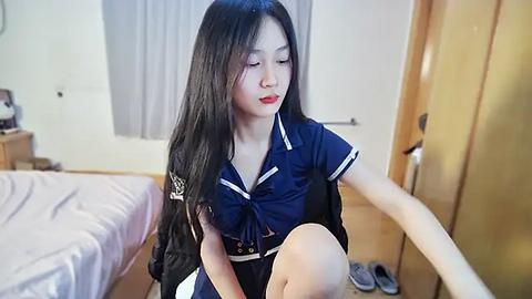 Media: Video of an East Asian woman with long black hair, wearing a blue sailor school uniform, sitting on a bed in a minimalist bedroom with wooden furniture and white walls.