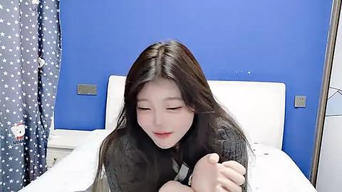 Media: Video of an Asian woman with long black hair, fair skin, and closed eyes, wearing a black sweater, lying on a white bed in a blue room with white walls and a patterned curtain.