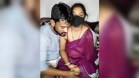 Media: Video of a South Asian couple, a man and a woman, sitting closely on a sofa. The woman wears a black face mask, a purple saree, and a gold necklace, while the man wears a white shirt and black mask.