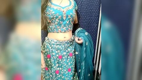 Media: Video of a woman wearing a vibrant blue, floral-patterned lehenga choli. She has long dark hair and a medium skin tone. Background is a dark blue textured wall.