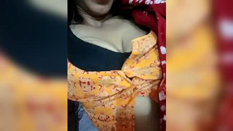 Media: Video of a woman with fair skin, wearing a yellow floral-patterned top, black bra, and a red polka-dot shirt, partially unbuttoned, showing cleavage. Background blurred.