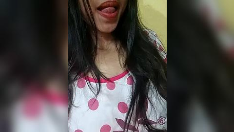 Media: Video of a young woman with long, straight black hair, wearing a white shirt with pink polka dots and a pink collar, playfully sticking her tongue out.