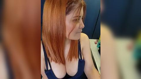 Media: A video of a fair-skinned woman with long, straight red hair and a septum piercing, wearing a low-cut blue tank top, sitting indoors.