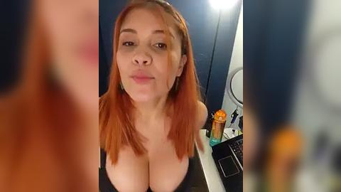 Media: Video of a light-skinned, red-haired woman with large breasts, wearing a black top, in a bathroom setting with a blue wall, sink, and orange soap dispenser.