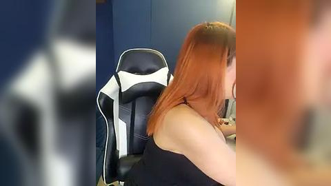 Media: Video of a woman with shoulder-length auburn hair, wearing a black sleeveless top, seated at a gaming chair with white and black accents in a dimly lit room.