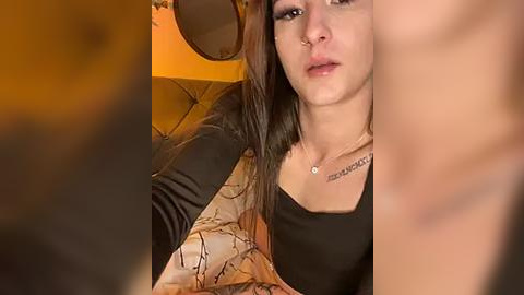 Media: A video shows a young woman with long, straight brown hair, light skin, and a nose ring, wearing a black top. She is in a dimly lit room, with a blurred, tan leather couch and a wooden chair in the background.