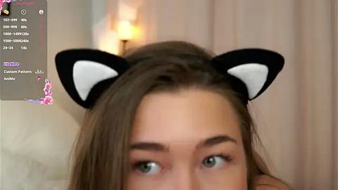 Media: Video of a young woman with light skin and green eyes, wearing a black and white cat ear headband. Background shows a blurred, beige curtain and a lamp.