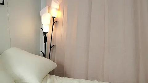 Media: Video of a minimalist bedroom corner with a white bed, beige curtains, and a tall, modern floor lamp with three white shades casting a warm glow.