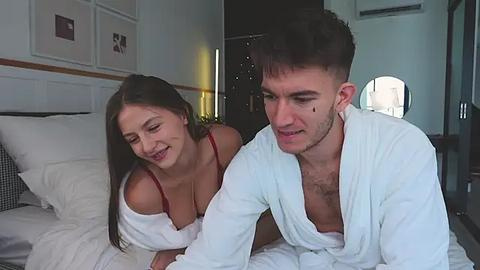 Media: Video of a smiling, topless woman in a red bra and a man in a white robe, both on a bed in a modern hotel room with white walls and a large mirror.