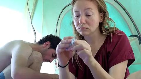 Media: Video of a shirtless young man with short dark hair lying on a bed, eyes closed, and a young woman with long blonde hair, wearing a red top, holding a ring, looking focused, in a tent with a turquoise background.