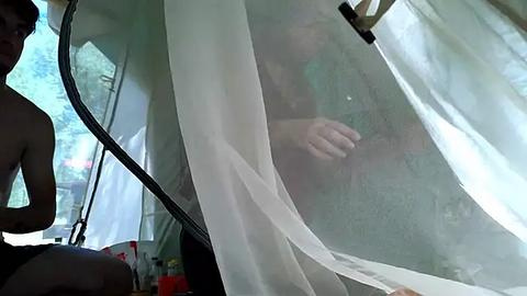 Media: Video of a semi-transparent tent with a person inside wearing a green shirt, partially visible through the fabric, in a forested outdoor setting.