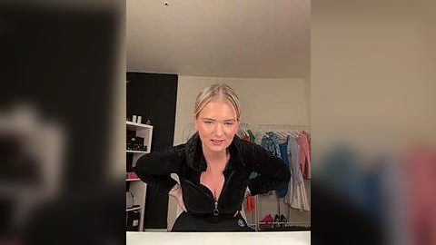 Media: Video of a blonde woman in a black zip-up jacket, leaning over a counter in a cluttered, beige-walled room with hanging clothes.