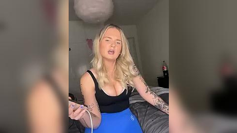 Media: Video of a blonde woman with tattoos, wearing a black tank top and blue leggings, holding a phone, in a dimly lit bedroom with a bed and white walls.