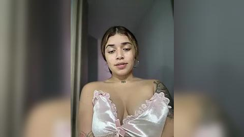 Media: A video of a young woman with light skin and short, dark hair, wearing a pink satin bra with lace trim, standing indoors against a gray wall.