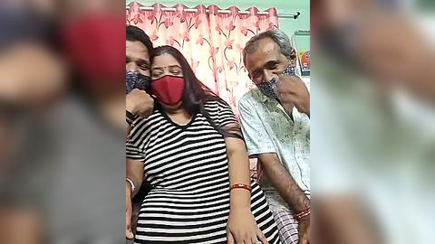 Media: Video of three people wearing face masks, a woman with long hair and a striped shirt, an elderly man in a checkered shirt, and a man with a beard, standing indoors against a pink floral curtain.