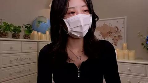 Media: Video of an Asian woman with long black hair, wearing a white mask, black top, and necklace, in a softly lit room with candles, plants, and a map on a white dresser.