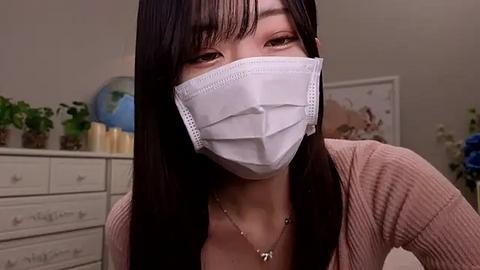 Media: Video of an East Asian woman with long black hair and bangs, wearing a white surgical mask and a beige ribbed sweater. She has a delicate necklace. Background features a white dresser with potted plants and a globe.