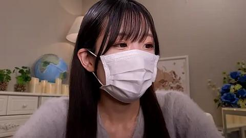 Media: Video of an Asian woman with long, straight black hair and fair skin, wearing a white surgical mask, seated indoors. Background includes a white dresser, candles, globe, and blue flowers.