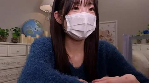 Media: Video of a young Asian woman with long black hair, wearing a white mask and blue fuzzy sweater, sitting indoors with a white dresser, candles, and a globe in the background.