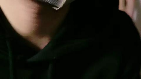 Media: Close-up video of a person's neck and upper chest, wearing a black shirt, with a grey cloth mask partially covering their mouth and nose, set against a dark background.