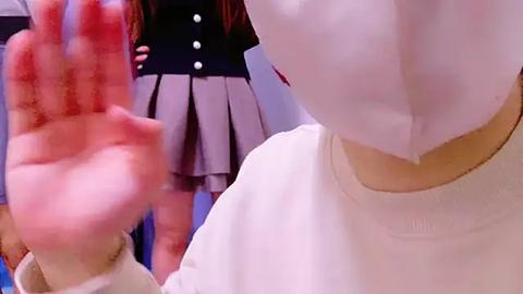 Media: Video of a person wearing a white surgical mask, with a blurred figure in a school uniform standing in the background. The image captures a moment of interaction or observation.