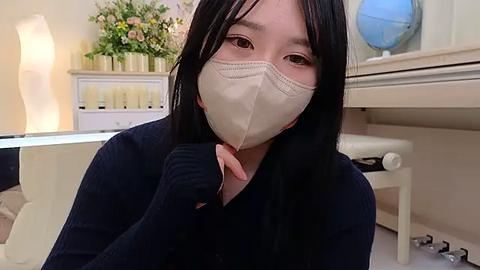 Media: Video of an East Asian woman with long black hair and pale skin wearing a beige surgical mask, navy sweater, and seated in a clinical setting with white furniture, flowers, and medical equipment.