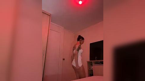 Media: A video of a woman in a white tank top and red bra, holding a knife, standing in a dimly lit, red-lit bedroom with a closed door and TV.