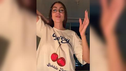 Media: A video of a young woman with fair skin, wearing glasses, a white T-shirt with cherry pendant, and a \"Candy\" logo, in a dimly lit room.