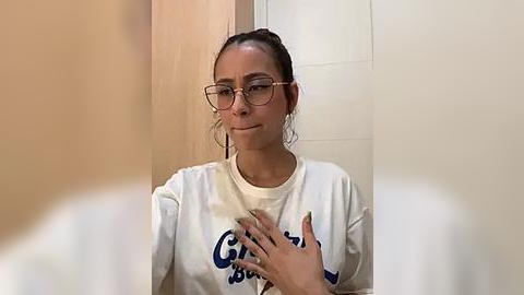 Media: Video of a young woman with medium skin tone and dark hair in a ponytail, wearing glasses and a white shirt, holding her chest in a bathroom with beige tiles and wooden door.