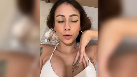 Media: A close-up video of a Latina woman with light brown skin, dark hair, and medium-sized breasts, wearing a white bikini top. She has a tattoo on her chest and is lying on a bed, eyes closed, with a serene expression.