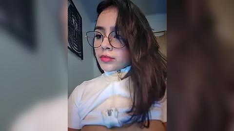 Media: Video of a young woman with light brown skin, long dark hair, and glasses, wearing a white crop top that reveals her midriff, taken indoors.