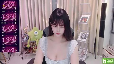 Media: A video of an East Asian woman with black hair, wearing a lacy white top, sitting in a modern, beige room. Background includes a wall-mounted TV, plush toys, and a framed photo.