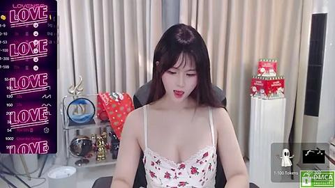 Media: Video of an East Asian woman with long black hair, wearing a white floral lingerie top, sitting in a modern office. Background includes a black desk with a computer, a calendar, and Christmas decorations.