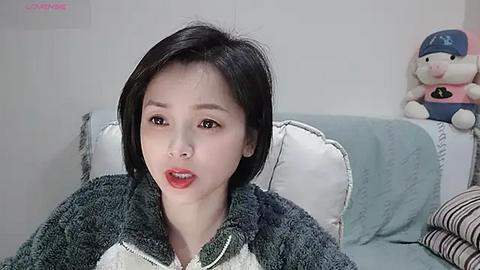 Media: Video of a young East Asian woman with short black hair, wearing a green zip-up hoodie, sitting on a light blue couch. A plush toy is beside her.