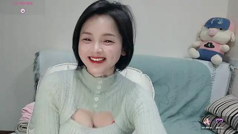 Media: A video of an Asian woman with fair skin, short black hair, and a cheerful smile, wearing a light green, textured sweater with a deep cutout revealing cleavage. She sits on a light blue couch with stuffed toys, including a Pikachu, in the background.