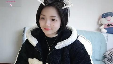 Media: Video of an Asian woman with short black hair, wearing a black and white fuzzy coat, white reindeer antlers, and a black top, smiling softly, in a cozy room with a blue cushion and stuffed toy in the background.