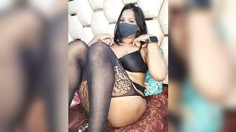 Media: Video of a woman with medium skin tone, wearing black lace lingerie, thigh-high stockings, and a face mask, sitting on a patterned bedspread.