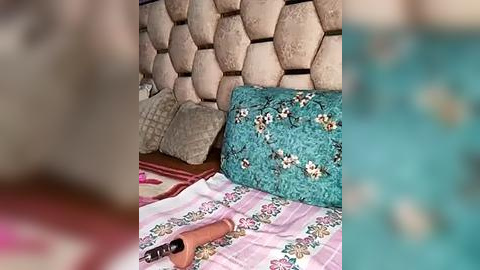 Media: A video shows a bed with a textured beige headboard and a turquoise pillow adorned with floral embroidery. The bedspread features pink and white floral patterns. A wooden-handled knife is placed on the bed.