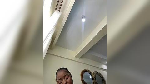 Media: Video of a young African-American girl with short hair, smiling in a modern, bright room with beige walls, white ceiling beams, a round mirror, and a white light fixture.