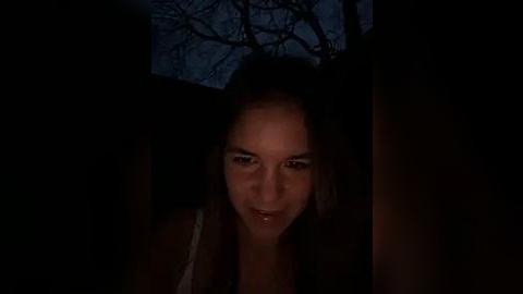 Media: Video of a young woman with long, dark hair, fair skin, and brown eyes, partially hidden in a dimly lit, narrow space. The background shows bare tree branches against a dark, night sky.