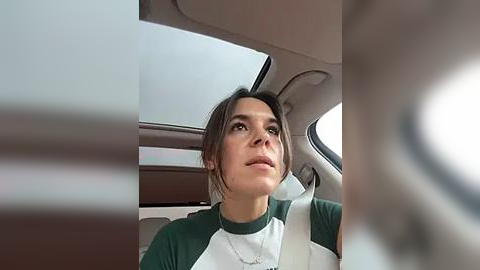 Media: Video of a woman with medium skin tone and brown hair in a ponytail, wearing a green and white t-shirt, driving a car, with the car's interior visible, including a white seatbelt and a beige dashboard.