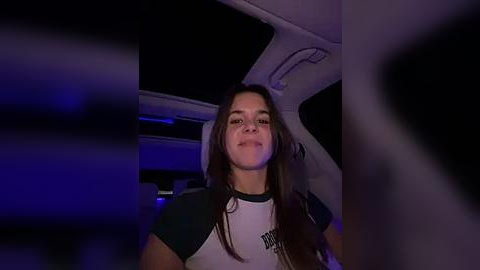 Media: Video of a young woman with long, dark hair, wearing a black and white T-shirt, sitting in a dimly lit car interior with purple ambient lighting.