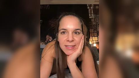 Media: Video of a smiling woman with long brown hair, lying on a bed, holding her chin. Background includes dimly lit room with patterned wall and candles.