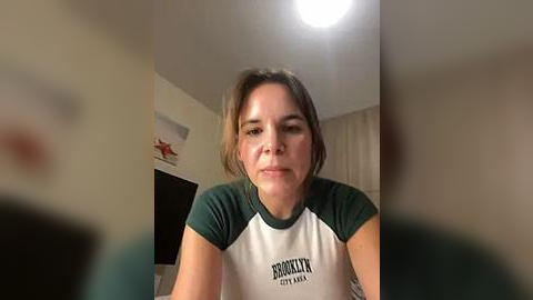 Media: Video of a woman with light brown hair, wearing a white and green t-shirt, sitting on a bed in a dimly lit room with beige walls and a hanging red starfish.